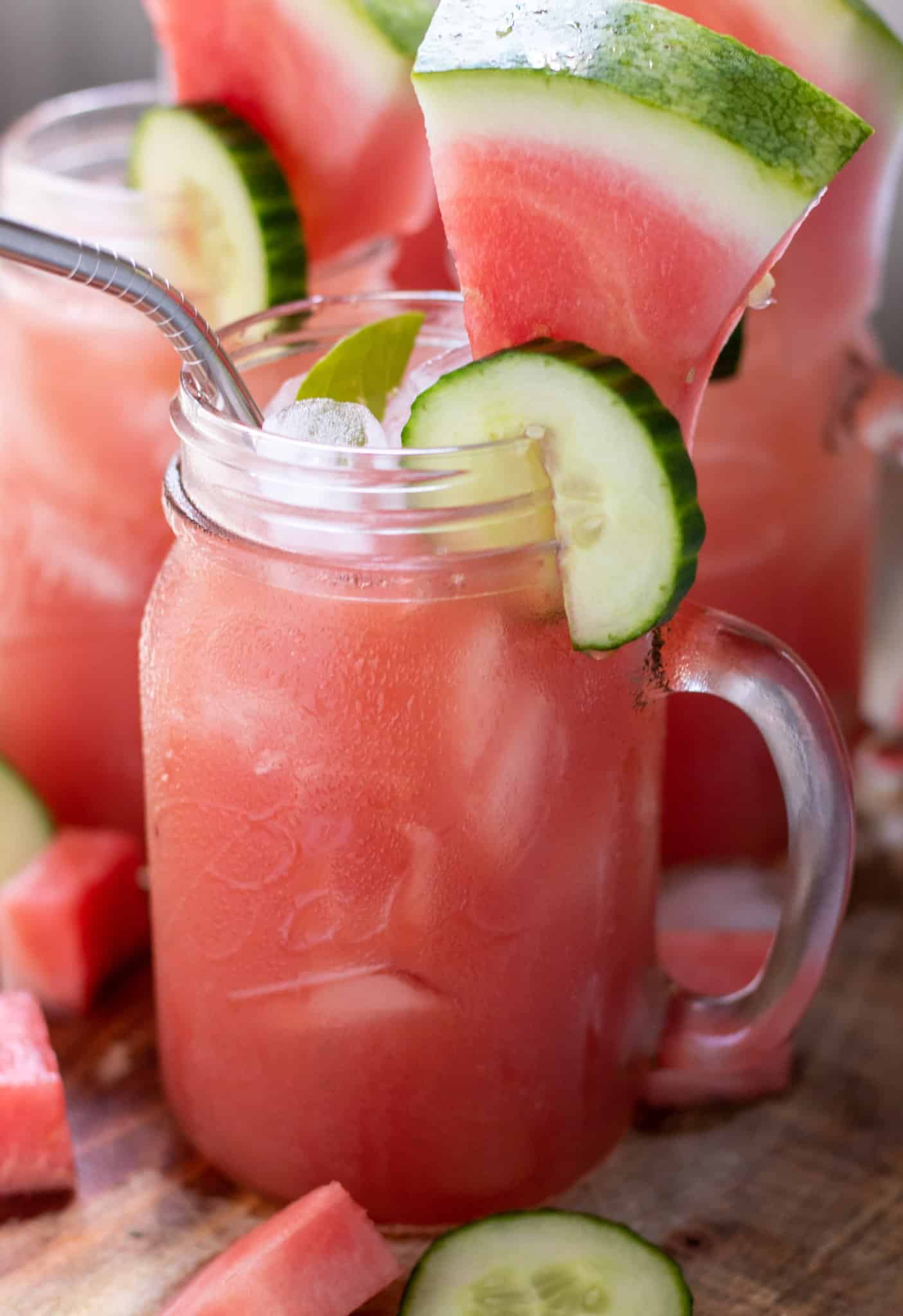 mason jar drink glasses filled with watermelon lemonade. They're garnished with a cucumber slice and small lemon wedge on the rim. The glasses have a metal drinking straw in them with ice in the glass.