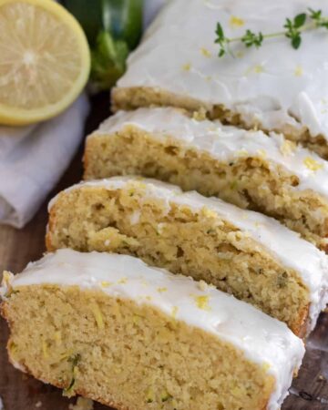 A wooden background with a loaf of lemon glazed zucchini bread that's sliced into 3 slices. There's a sprig of fresh thyme on top of the loaf with a half a lemon and zucchini in the background
