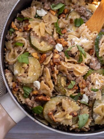 A large sauce pan filled with creamy pasta with zucchini, sausage and goat cheese. It's topped with toasted walnuts, goat cheese crumbles and chopped fresh basil