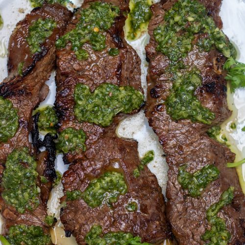 A white round dish topped with 3 grilled sirloin steaks that are drizzled with chimichurri sauce. There's fresh cilantro and a wedge of lime on the plate.
