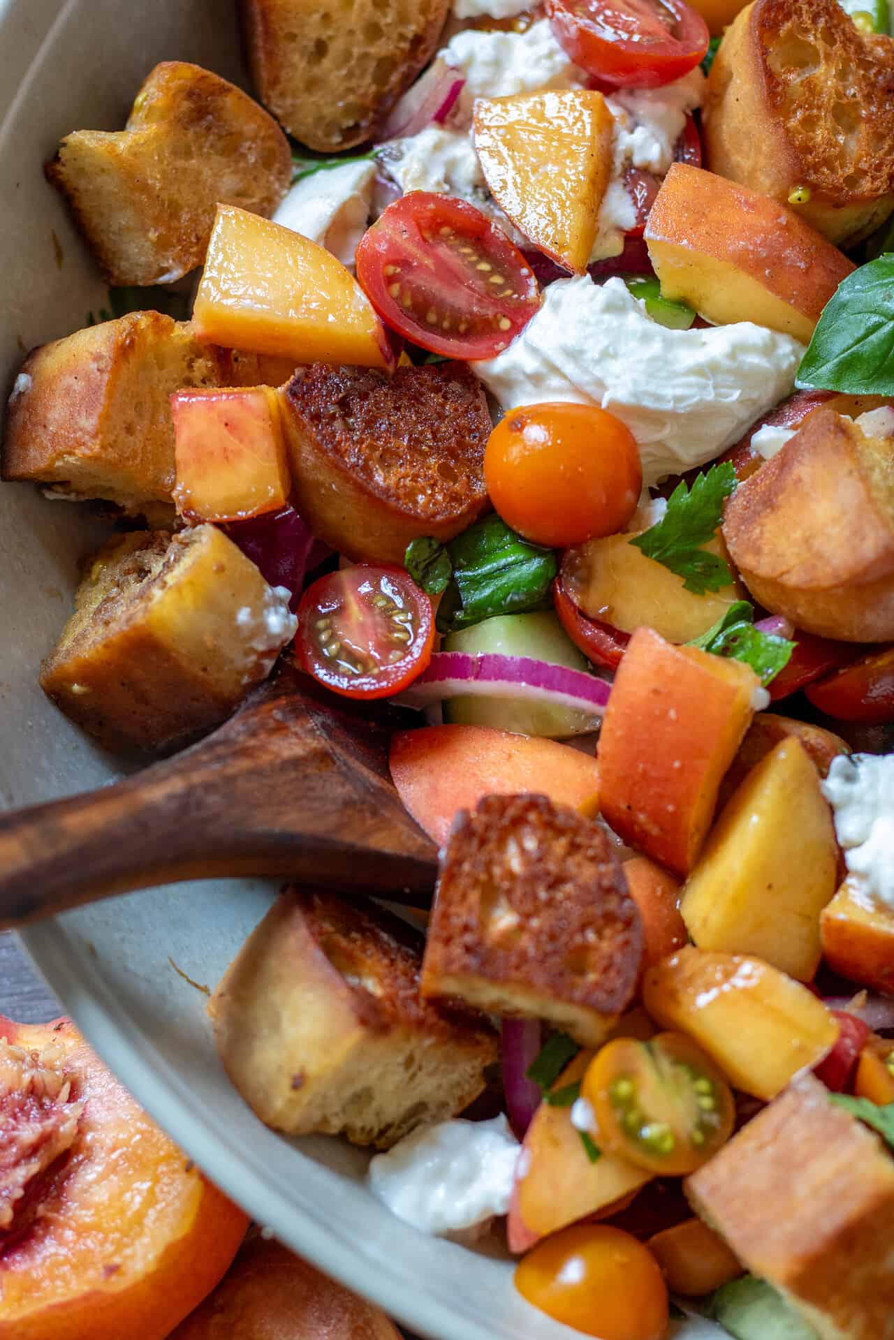 A large beige serving bowl filled with summer panzanella salad with peaches and burrata. There's a wooden serving spoon in the bowl and a halved peach next to the bowl.