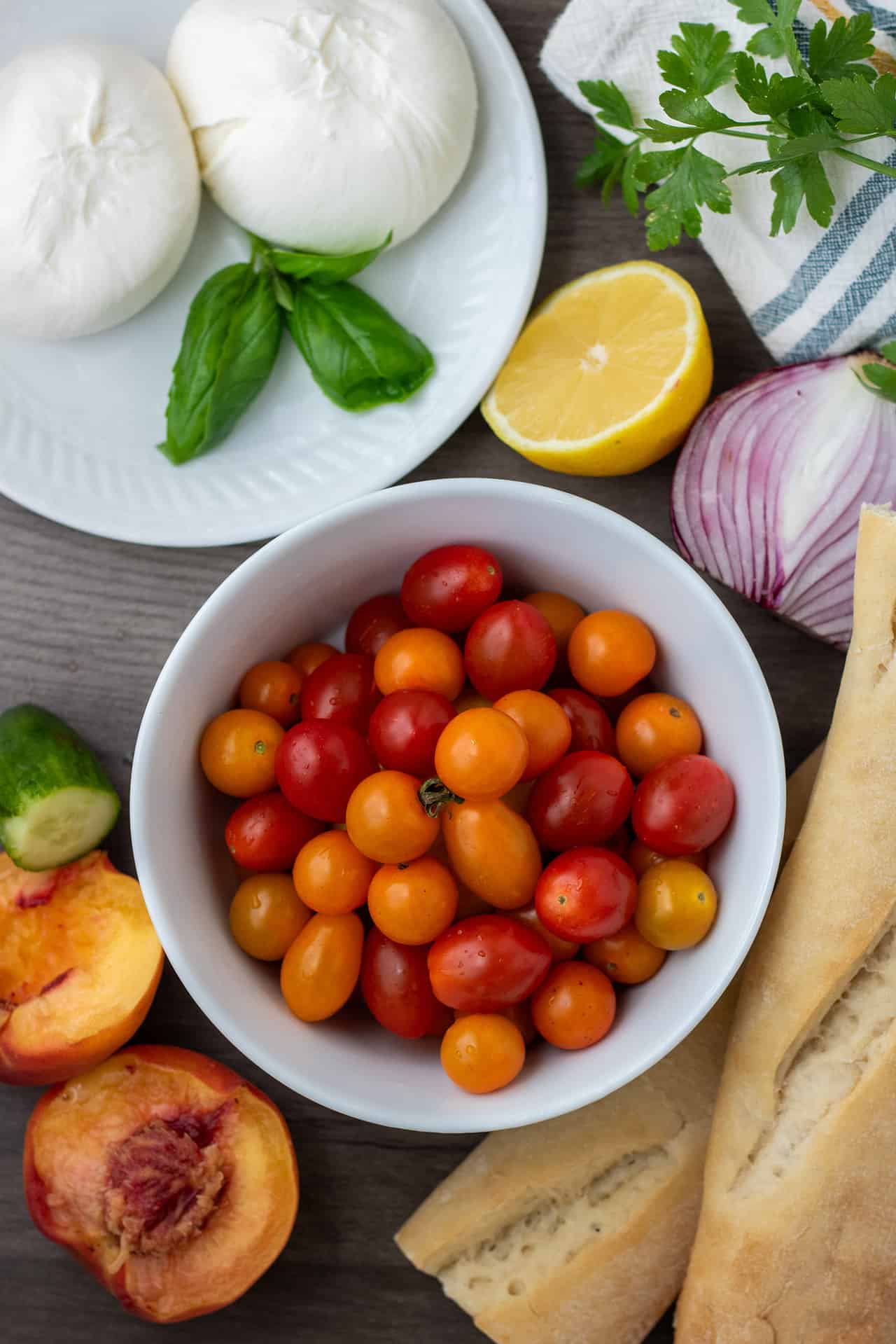 A white bowl filled with red and orange cherry tomatoes. There's a small white plate with burrata cheese and fresh basil. There's a half of a lemon and red onion in the background with a loaf of French bread.