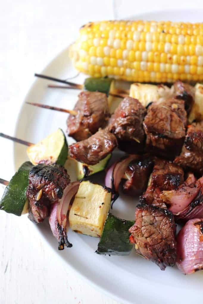 A white platter of steak skewers that have been marinated with bourbon brown sugar marinade.  There's a cob of corn in the background