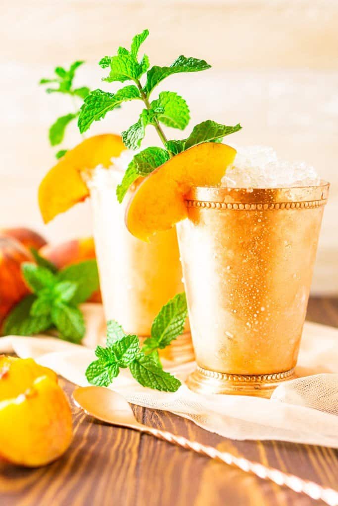 Two copper cups with peach mint julep.  The cups are ice cold and sweating.  There's a fresh peach slices and mint sprig for garnish.  There's fresh sliced peaches and extra mint in the background.