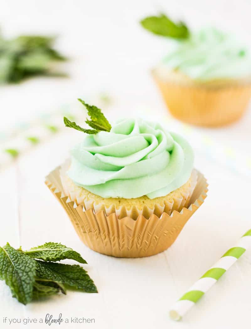 A mint julep cupcake with a gold cupcake liner and topped with fresh mint leaves.  There's a green and white striped straw, another cupcake and fresh mint leaves in the background