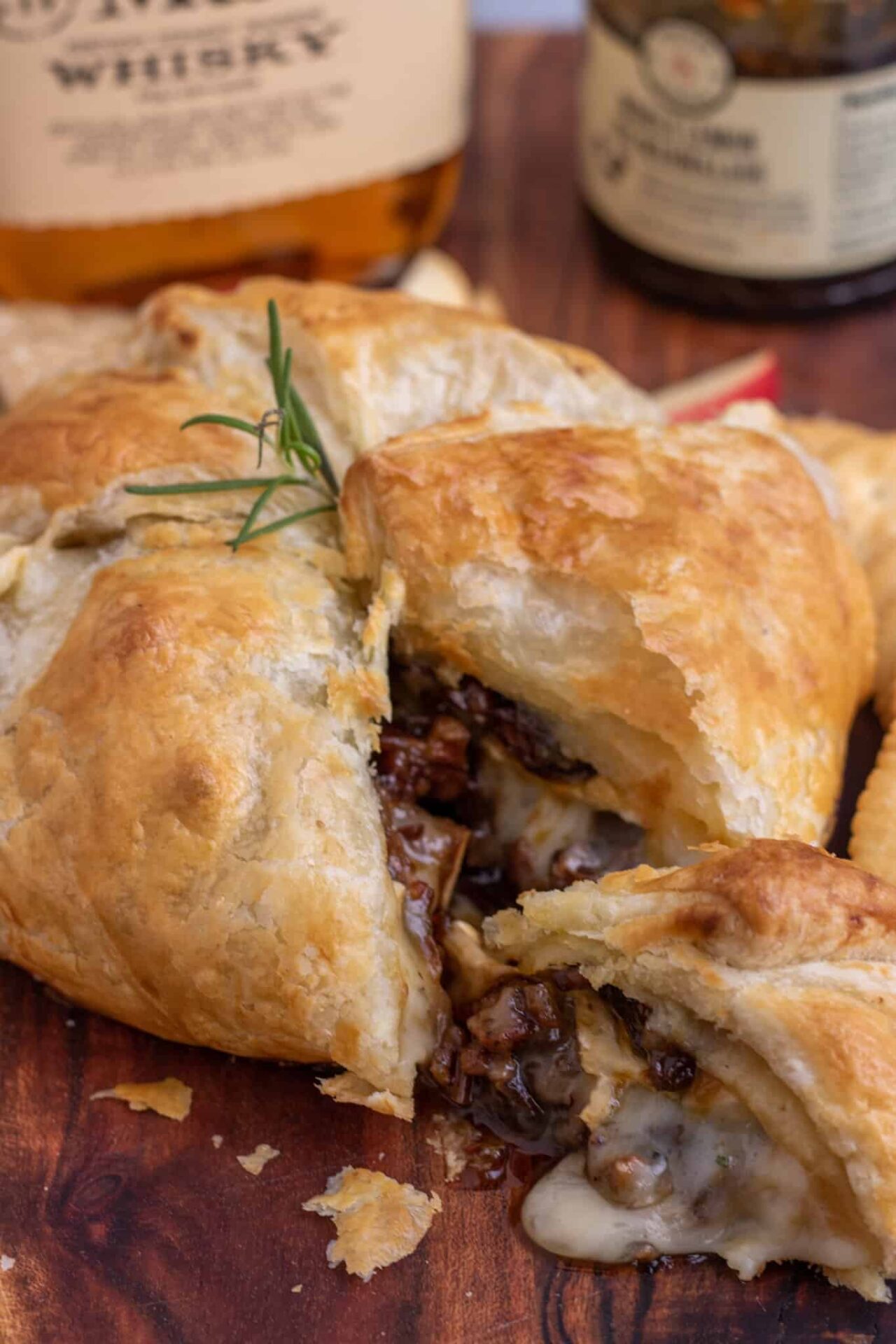 A wooden cutting board with a baked brie wrapped in puff pastry on top. The baked brie is cut into a wedge and there's melted cheese falling out of the side with pecans. It's topped with a sprig of rosemary and there's a bottle of whisky and jar of fig jam in the background