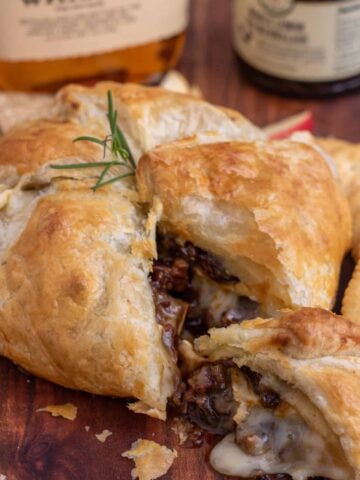 A wooden cutting board with a baked brie wrapped in puff pastry on top. The baked brie is cut into a wedge and there's melted cheese falling out of the side with pecans. It's topped with a sprig of rosemary and there's a bottle of whisky and jar of fig jam in the background