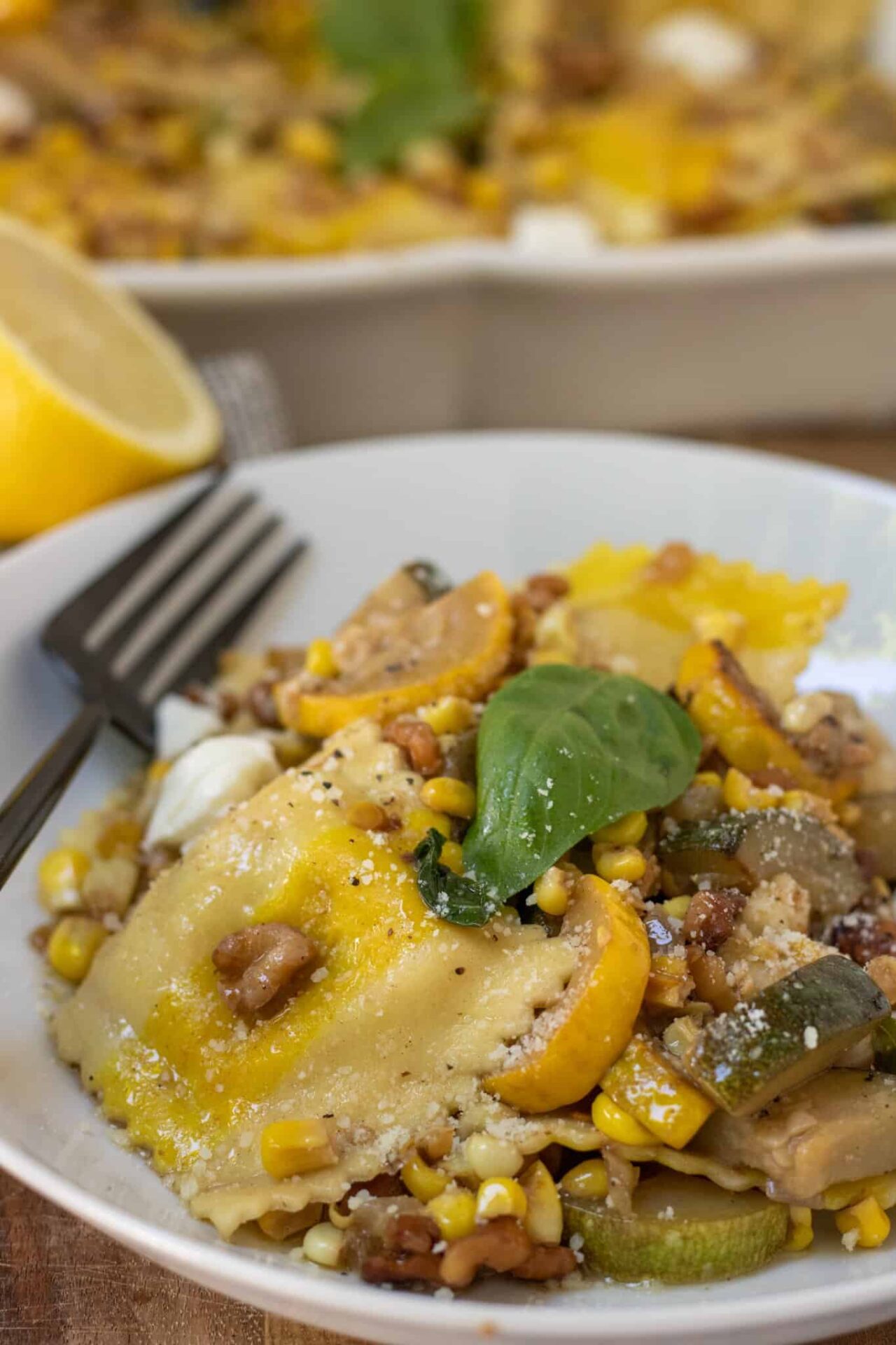 A white pasta bowl filled with yellow ravioli with sauteed summer squash, fresh basil, corn and melted pieces of mozzarella. There's half of a lemon in the background.