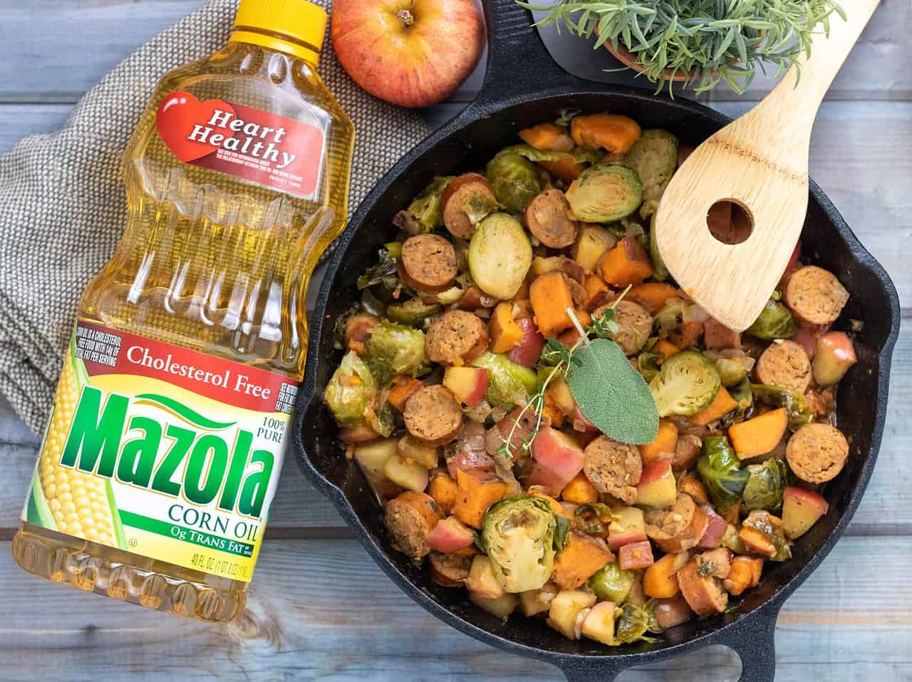 An overhead shot of a black cast iron pan filled with chicken sausage and fall vegetables. There's a wooden serving spoon halfway in the pan. To the left, there's a bottle of Mazola corn oil. Behind the skillet there's an apple and small rosemary plant.
