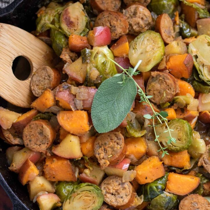 A cast iron skillet filled with chicken sausage, apple and veggies. It's got a wooden serving spoon in it and it's topped with a fresh sage leaf and thyme sprig