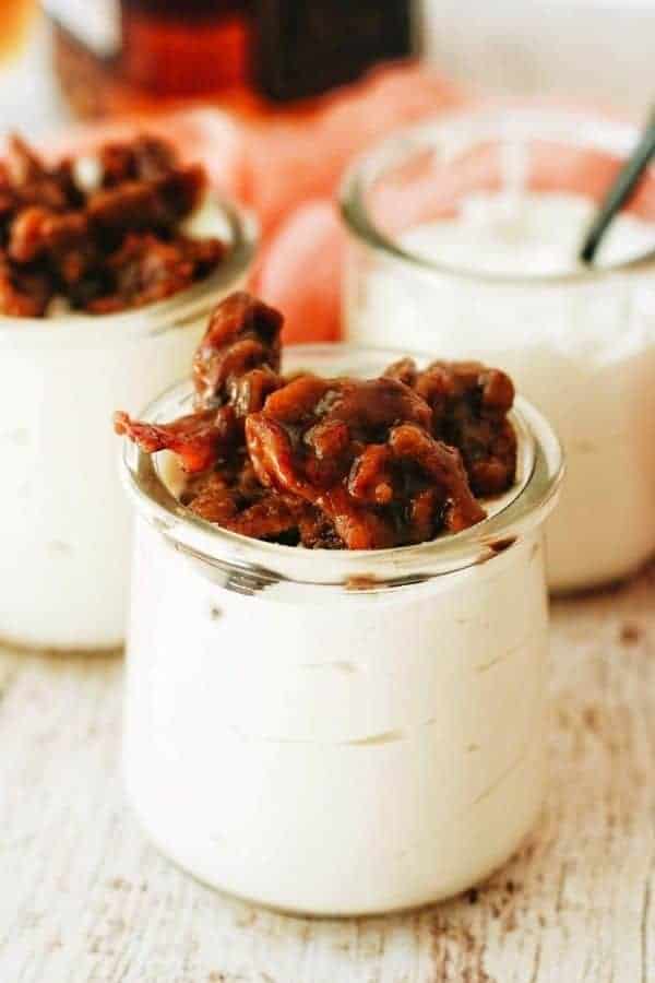 A small glass jar filled with no bake cheesecake that's topped with bourbon candied bacon.