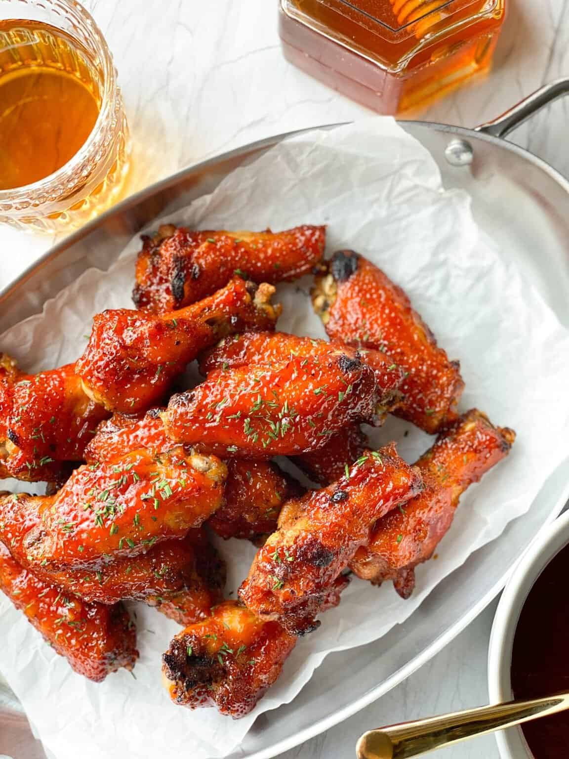 A serving platter piled with spicy honey bourbon wings. There's a side of honey in the background. The wings are on white parchment paper and they're crispy with a sweet and spicy glaze
