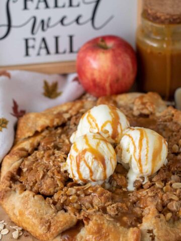 Apple crostata topped with 3 scoops of vanilla ice cream that's drizzled with caramel sauce. The crostata has an oat crumble and it's golden brown. There's a napkin with fall leaves next to it and a red apple with a jar of caramel sauce in the background