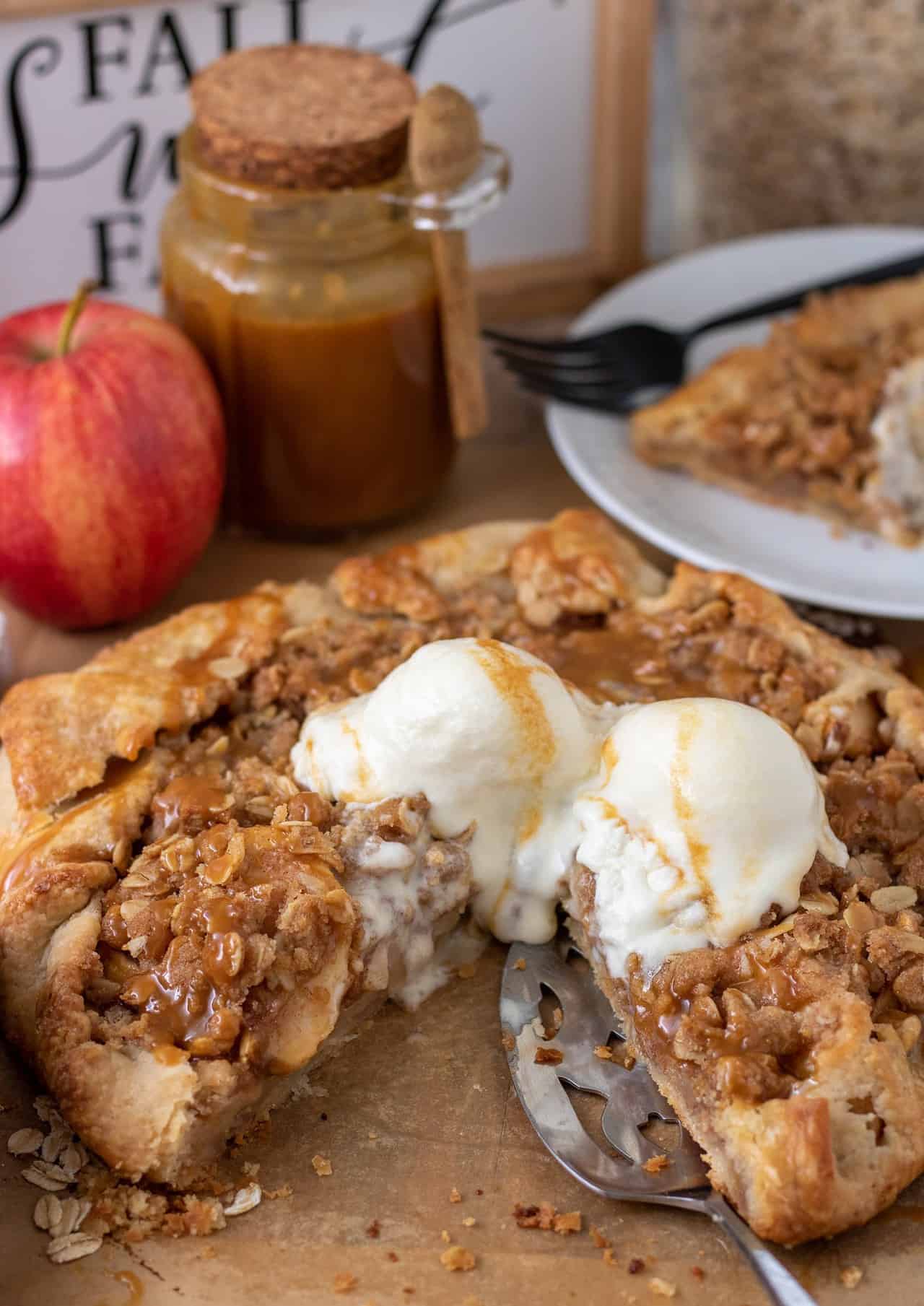 Apple crostata on brown parchment paper. There's a slice taken out of it with a pie server next to it. There's crumbs on the parchment paper. Two scoops of vanilla ice cream with caramel drizzle are in the middle of the crostata. There's a red apple and a jar of caramel sauce in the background next to a small white place with a slice of the crostata and a black fork.