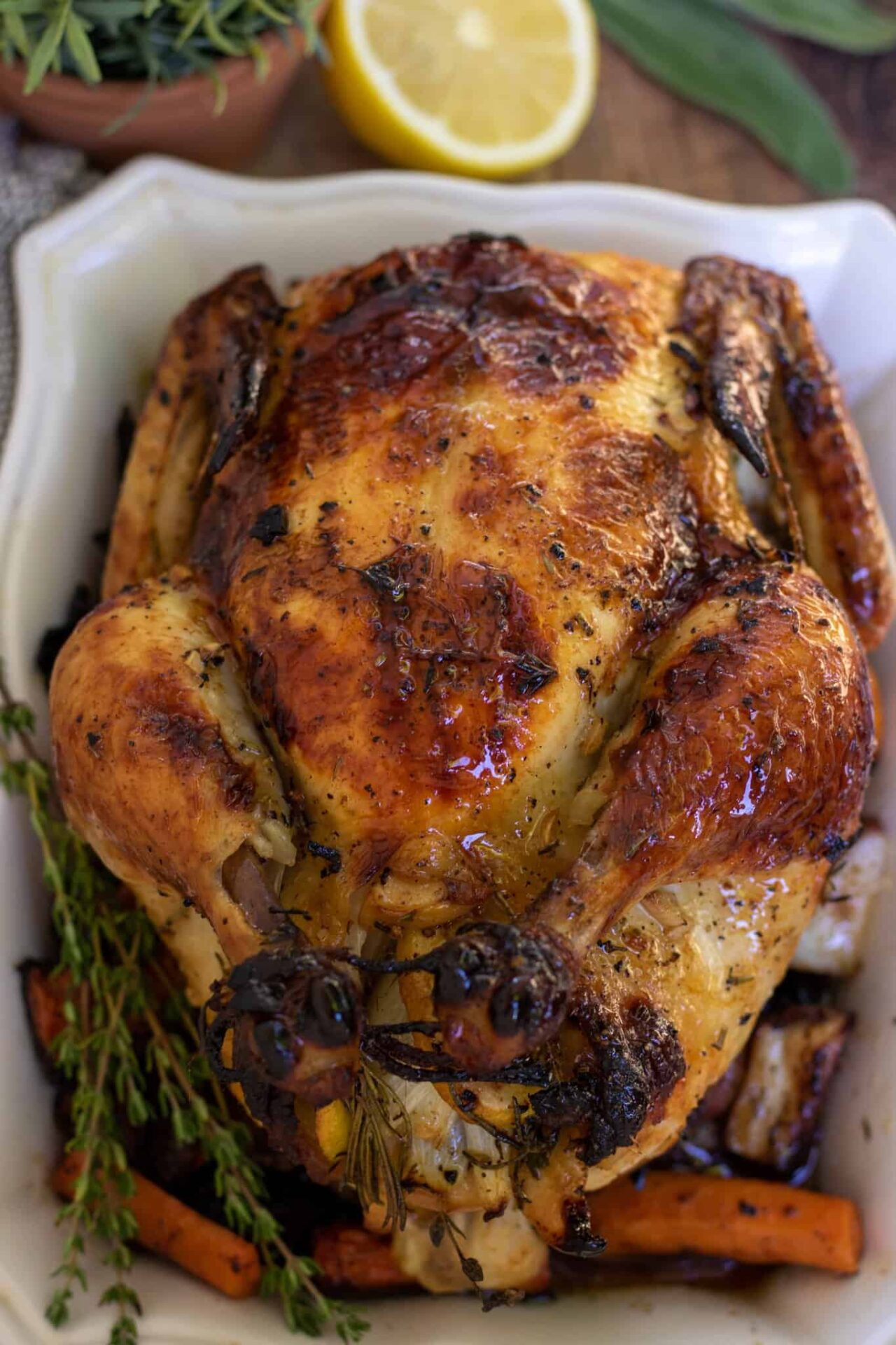 A whole roasted chicken on square white serving dish. The legs are tied up and the skin is browned and golden. There's a bunch of fresh rosemary and thyme next to the chicken and it's sitting on top of carrots, potatoes and onions.