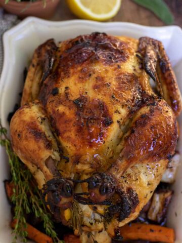 A whole roasted chicken on square white serving dish. The legs are tied up and the skin is browned and golden. There's a bunch of fresh rosemary and thyme next to the chicken and it's sitting on top of carrots, potatoes and onions.