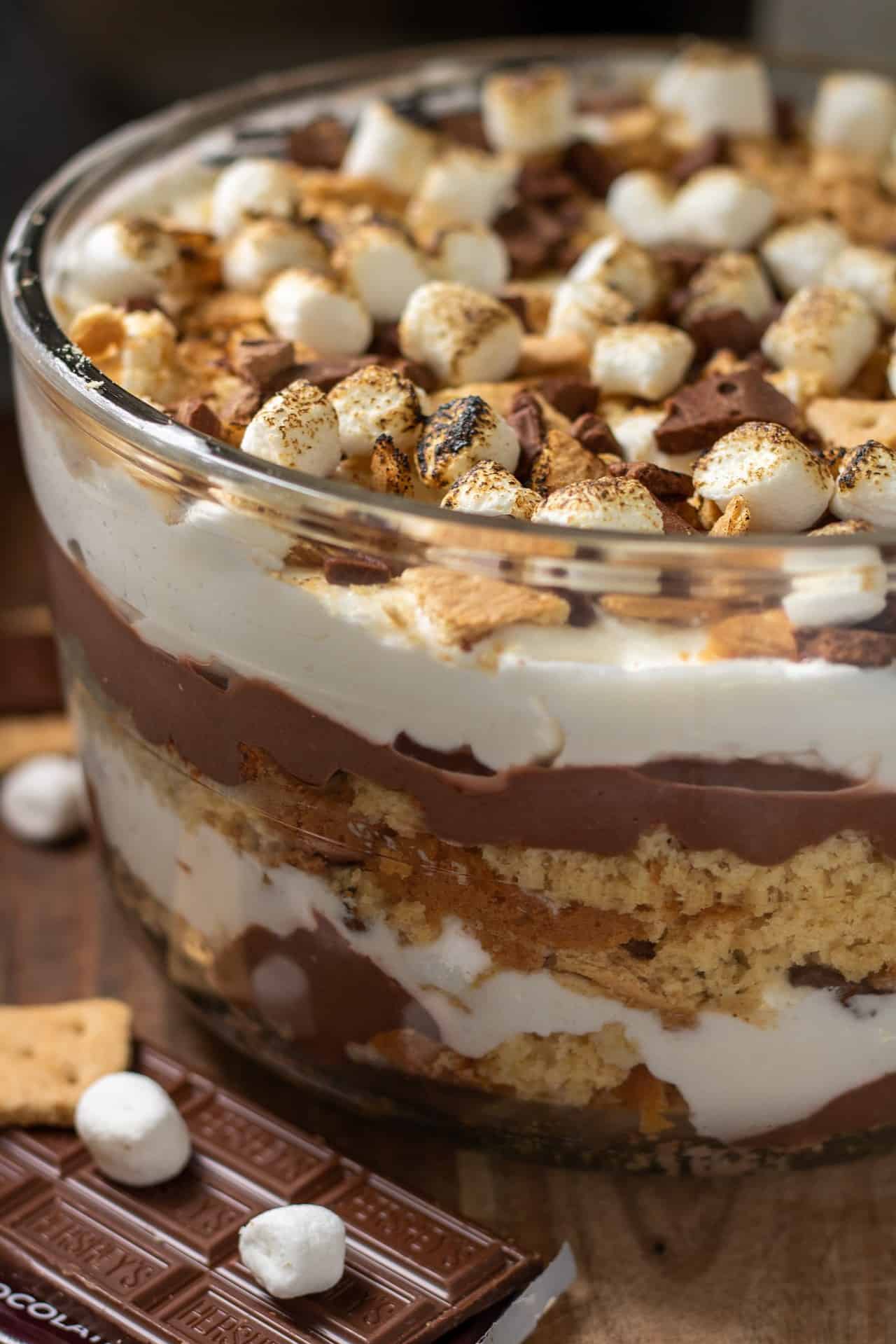 A large glass bowl on top of a wooden surface. The bowl is layered with yellow cake, chocolate pudding and whipped cream. It's topped with toasted mini marshmallows, graham cracker pieces and hershey's chocolate pieces There's a half of a hershey's chocolate bar next to it with some mini marshmallows and half of a graham cracker