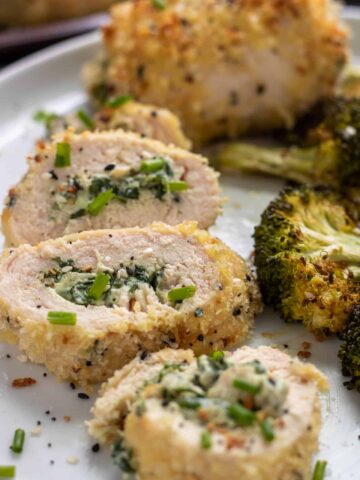 A white dish topped with sliced cream cheese stuffed chicken that's coated in panko and everything seasoning. There's roasted broccoli in the background and the chicken is sprinkled with green scallions..
