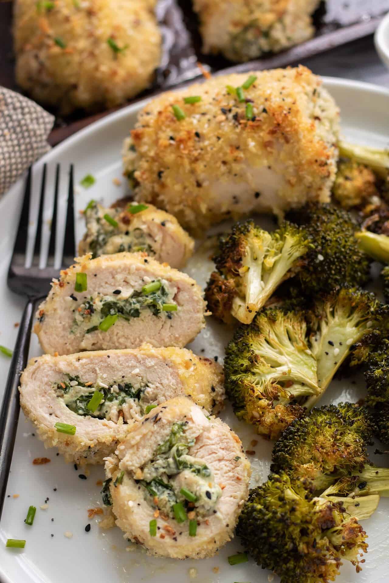 A white dish with sliced stuffed chicken that's been rolled up with cream cheese and spinach. There's roasted broccoli next to the plate with a whole baked chicken breast in the background. The chicken is coated in panko and it's golden. There's a black fork next to the chicken on the plate