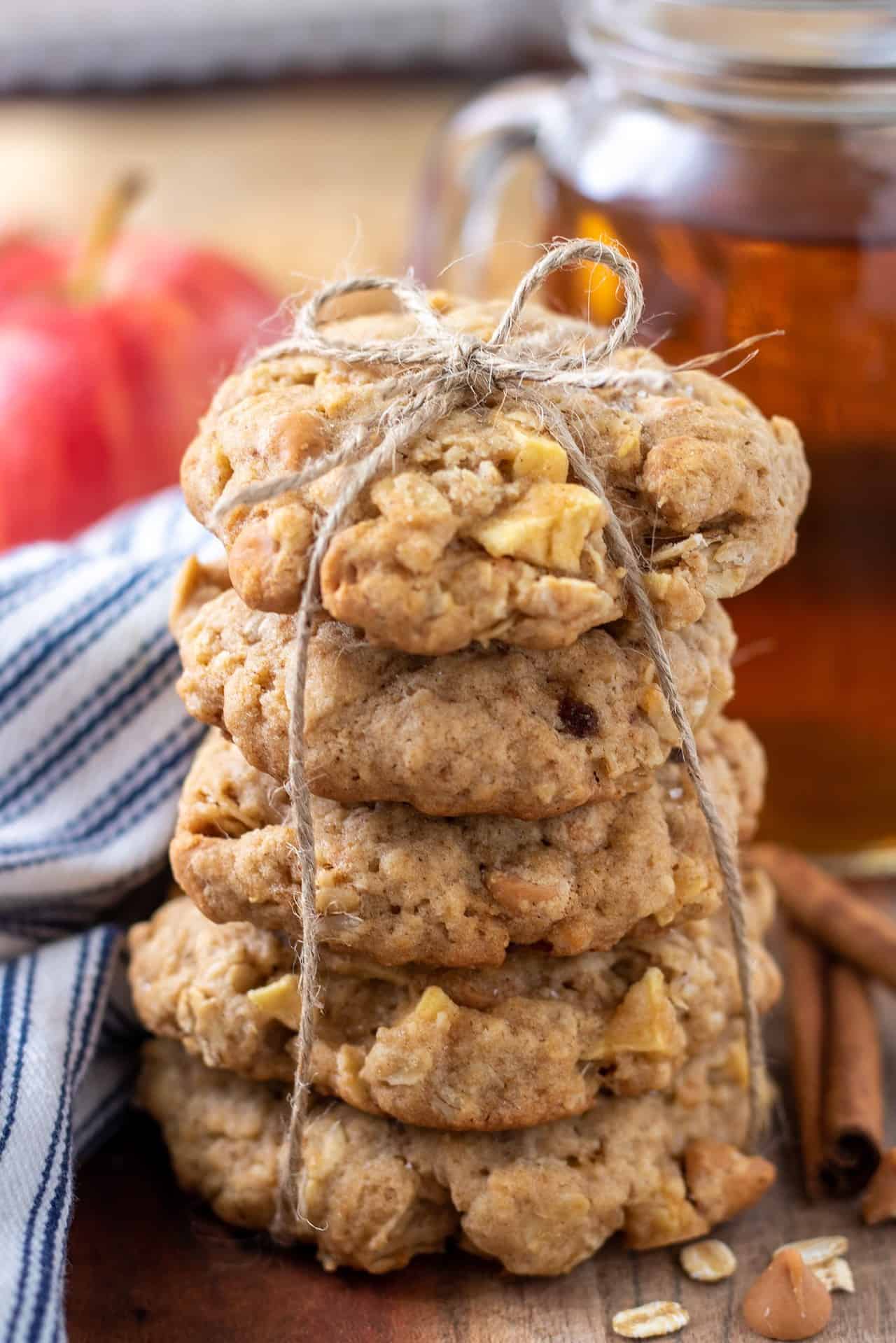 A stack of oatmeal cookies tied together with string. There's two cinnamon sticks next to it and a glass of hot apple cider in the background with a red apple