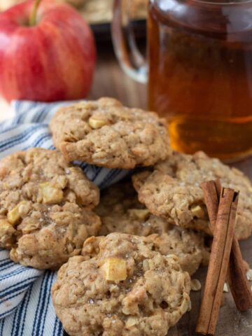 5 apple oatmeal cookies on a wooden surface. There's two cinnamon sticks next to the cookies. A red apple and a mason jar glass of hot apple cider are in the background
