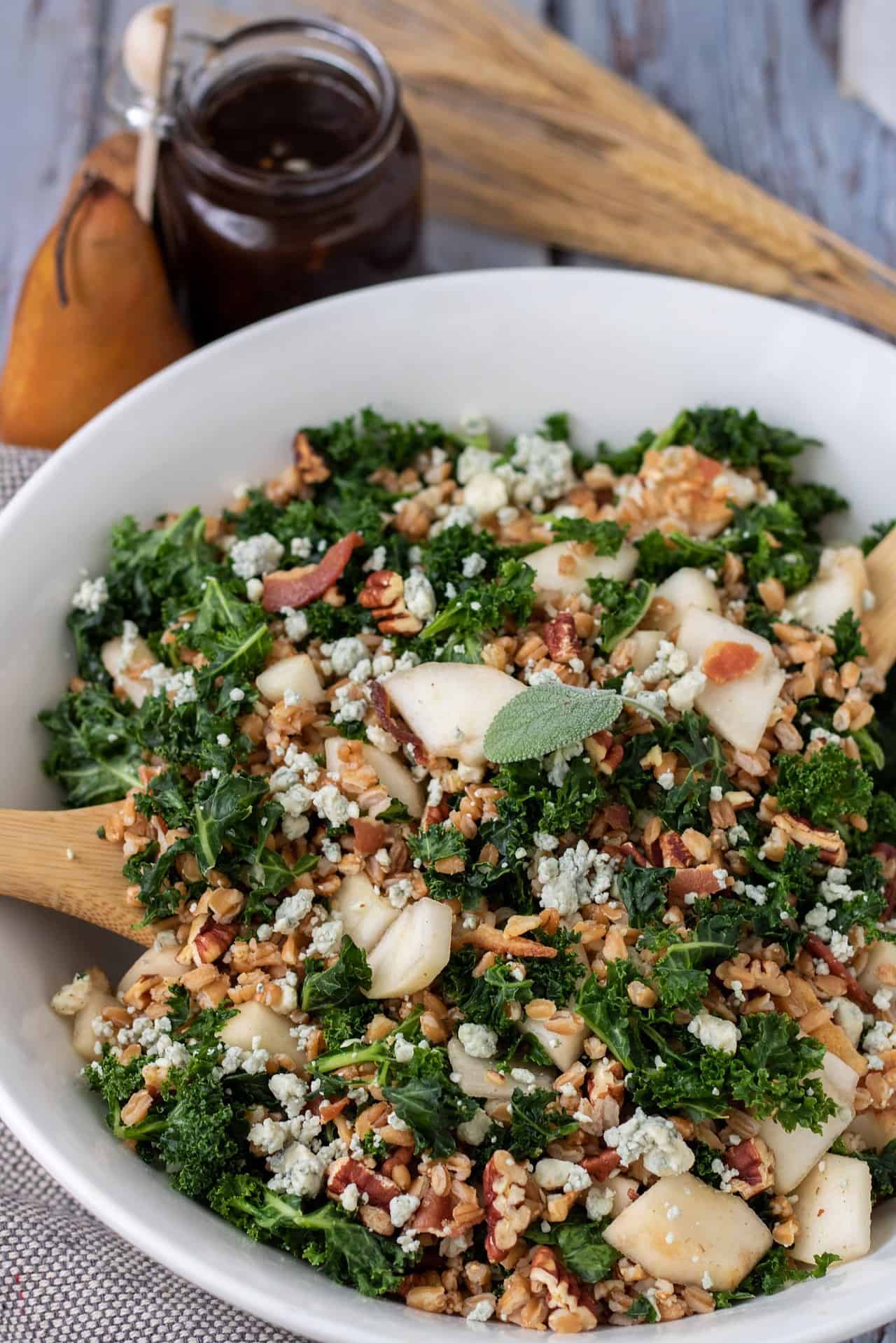 a large white serving bowl filled with kale and farro salad. It's got a fresh sage leaf on top and two wooden serving utensils in the salad. The small glass jar of balsamic fig dressing is in the background with a whole pear and dried wheat stalks.