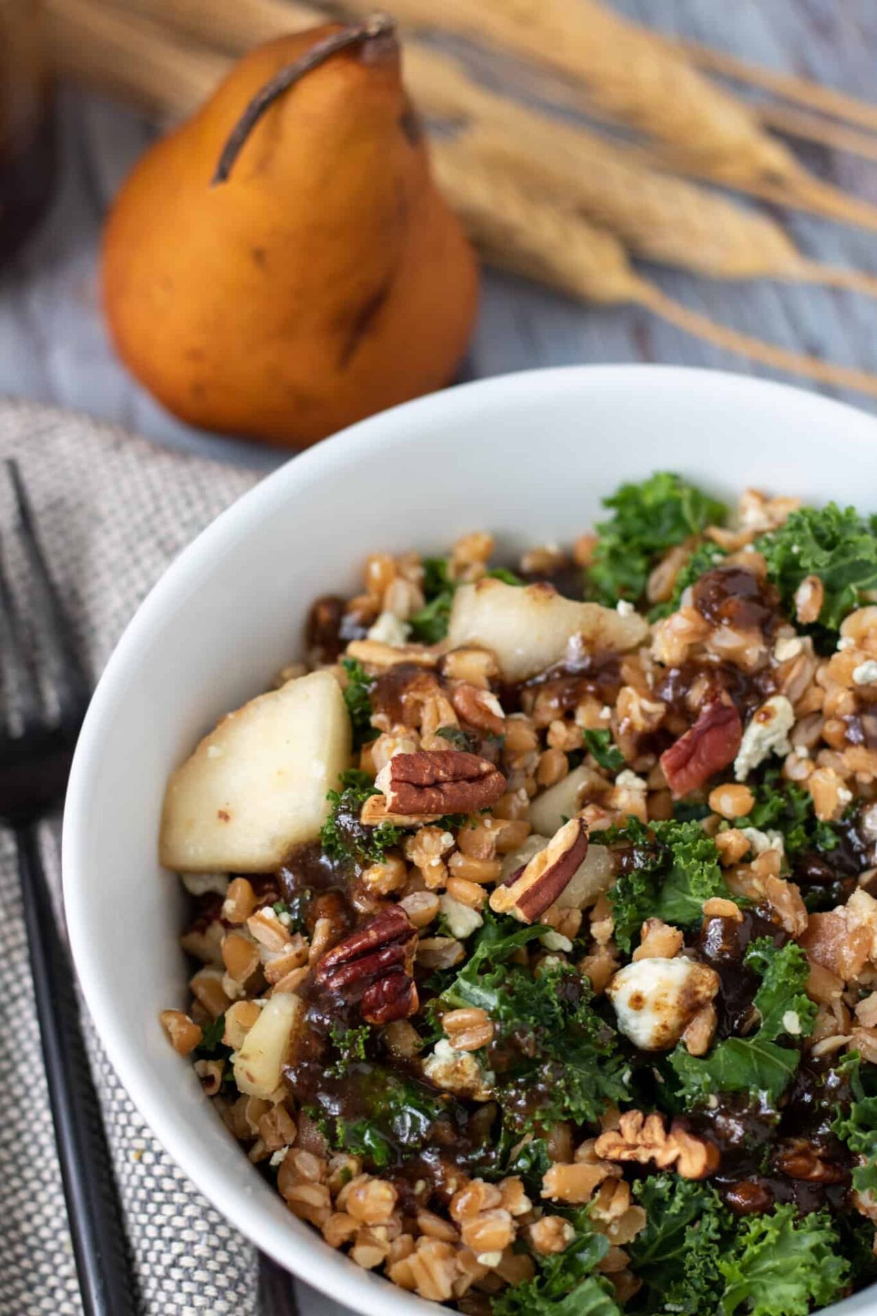 a white salad bowl with farro, kale, pecans, bacon, Gorgonzola crumbles and chunks of pears. There's a black fork next to the bowl. A whole pear is in the background with dried wheat stalks