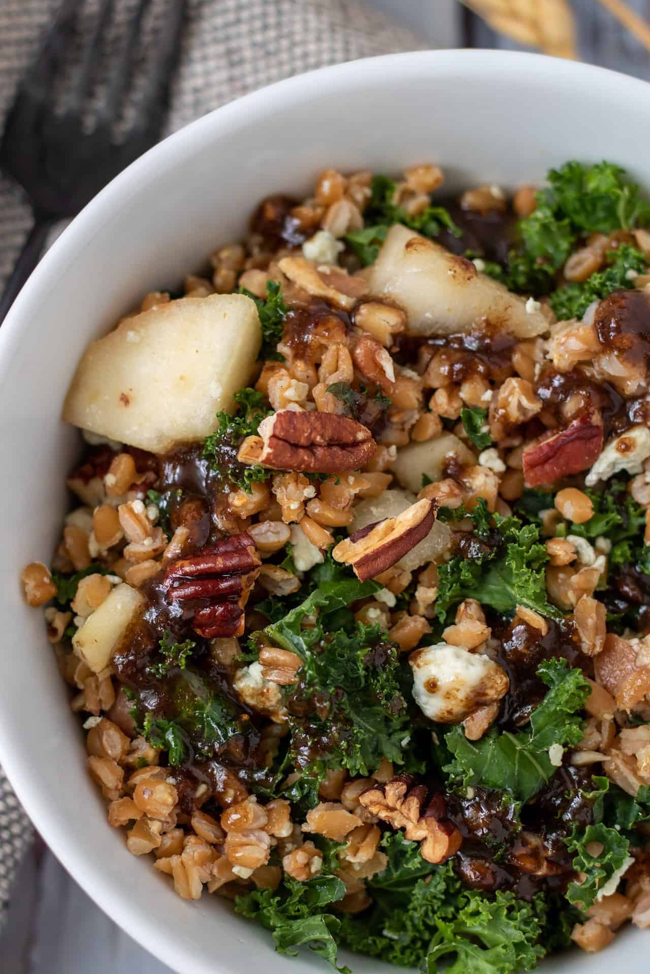 A white bowl filled with farro and kale salad with pecans, bacon, pears and Gorgonzola. There's a black fork next to the bowl.