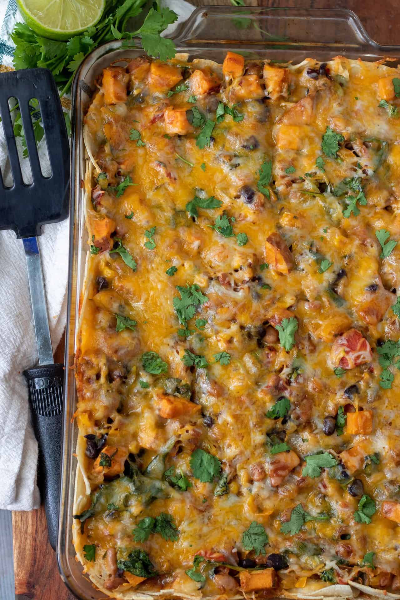 A glass baking dish with a vegetable enchilada casserole. It's topped with fresh cilantro and cheese. There's a spatula next to the baking dish for serving.