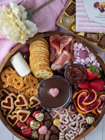 a charcuterie board filled with fruits, cheese, valentine's candies and chocolates. There's a white flower in the background with a box of chocolates