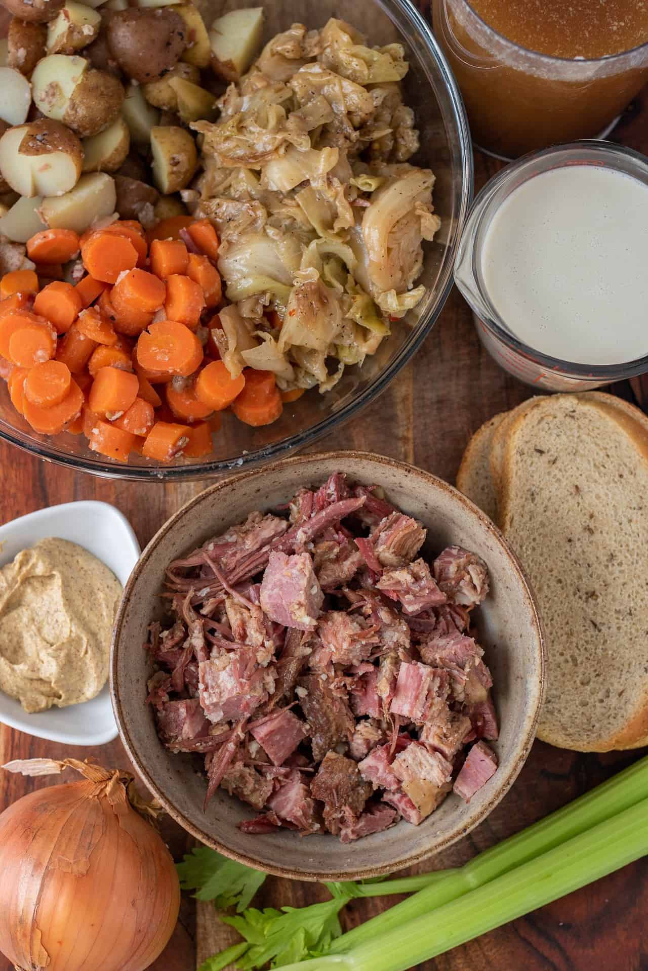 A wooden surface topped with corned beef soup ingredients. A bowl of cubed corned beef, a small dish of mustard, slices of rye bread, and a large bowl with sliced carrots, cabbage and potatoes. A measuring cup filled with milk is also on the wooden surface.