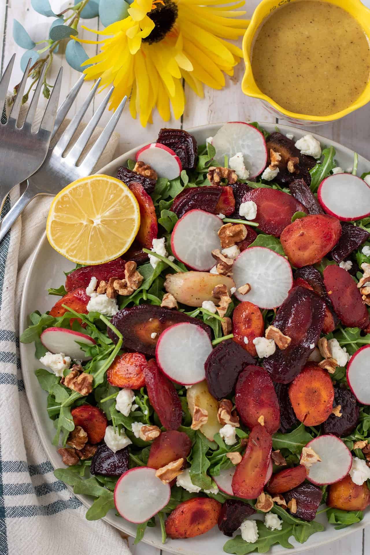 A large round serving plate with a roasted carrot, beet and arugula salad. The salad is colorful with sliced radishes, a halved lemon and goat cheese crumbles. Theres a silver serving fork next the plate and a large bright yellow flower.