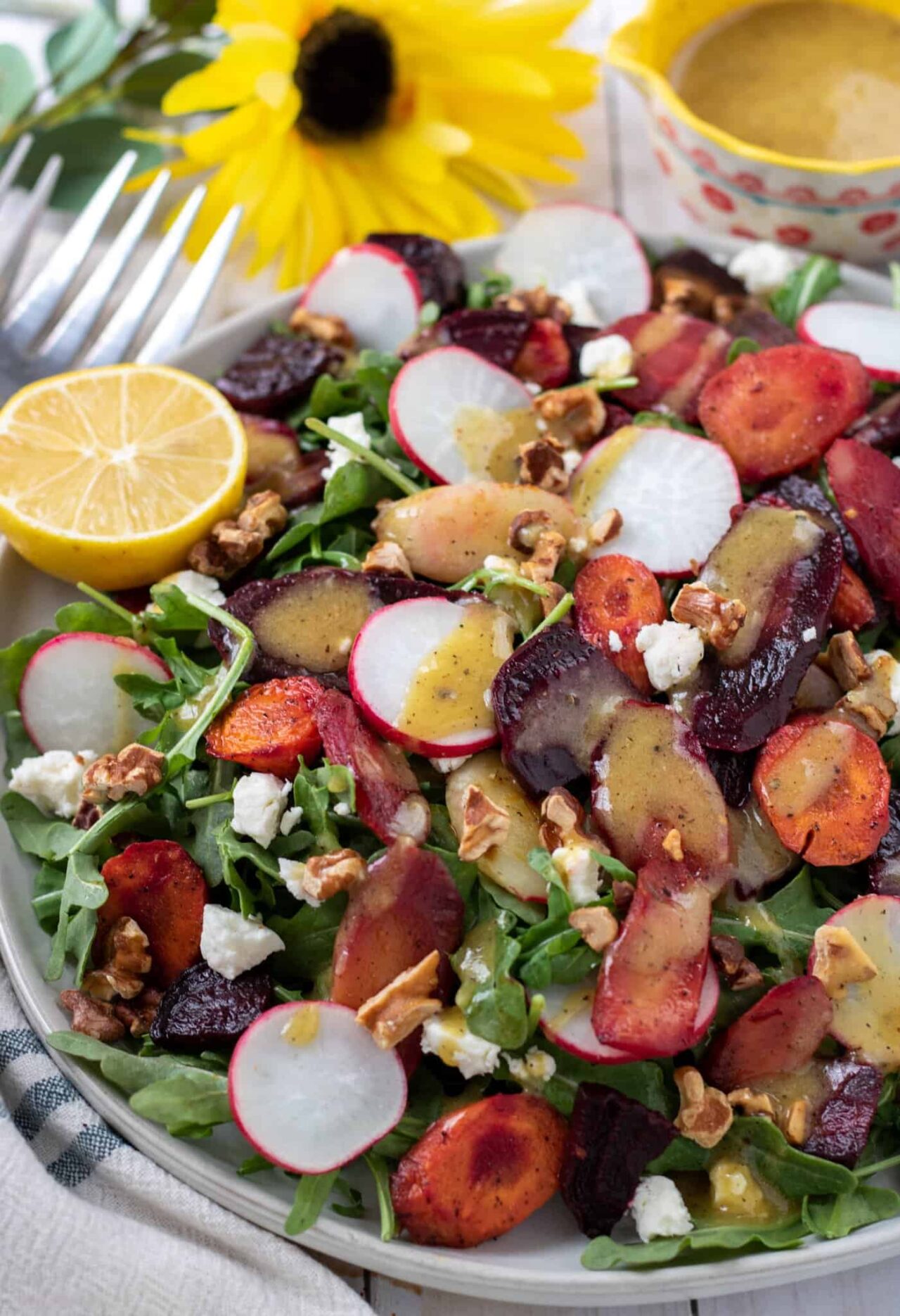 A large platter topped with a colorful arugula salad that has sliced radishes, goat cheese crumbles, toasted walnuts, roasted beets and rainbow carrots. There's a halved lemon on the side. A yellow daisy flower is in the background next to a small bowl of salad dressing.