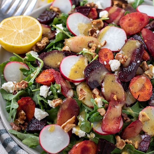 A large platter topped with a colorful arugula salad that has sliced radishes, goat cheese crumbles, toasted walnuts, roasted beets and rainbow carrots. There's a halved lemon on the side. A yellow daisy flower is in the background next to a small bowl of salad dressing.