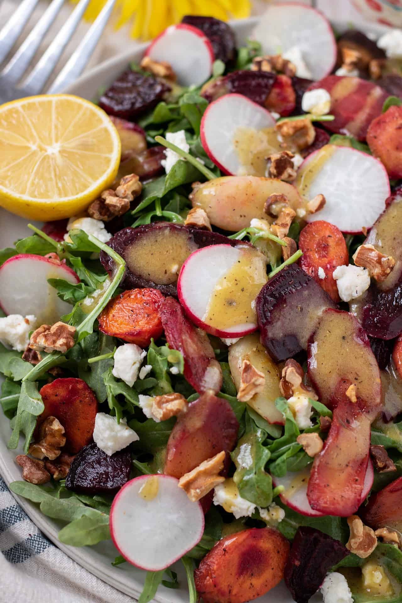 A close up shot of a large arugula salad that's topped with roasted rainbow carrots, beets, goat cheese, walnuts and radishes. There's a half of a lemon in the background and the salad is drizzled with a Dijon maple dressing.