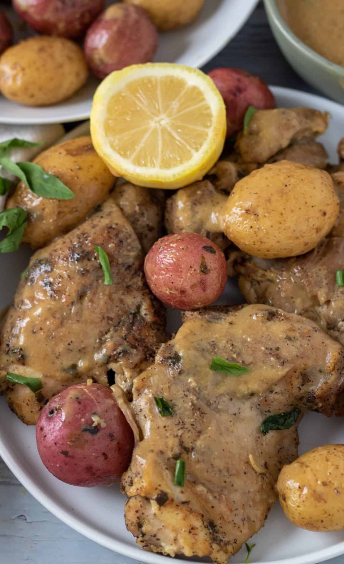 A white plate topped with chicken thighs with lemon Dijon gravy. There's fresh thyme sprinkled on the chicken and a half of lemon is sitting on the plate. There's yellow and red small potatoes.