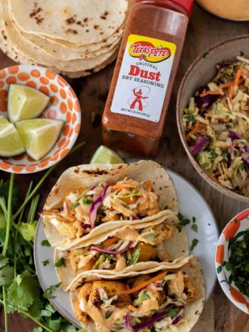 A wooden background with a small plate filled with color tacos. There's a small bowl of lime wedges, a bunch of fresh cilantro and grilled corn tortillas in the background. A container of seasoning is in the middle and you can see a bowl of colorful slaw next to the tacos