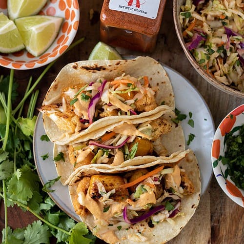 A wooden background with a small plate filled with color tacos. There's a small bowl of lime wedges, a bunch of fresh cilantro and grilled corn tortillas in the background. A container of seasoning is in the middle and you can see a bowl of colorful slaw next to the tacos