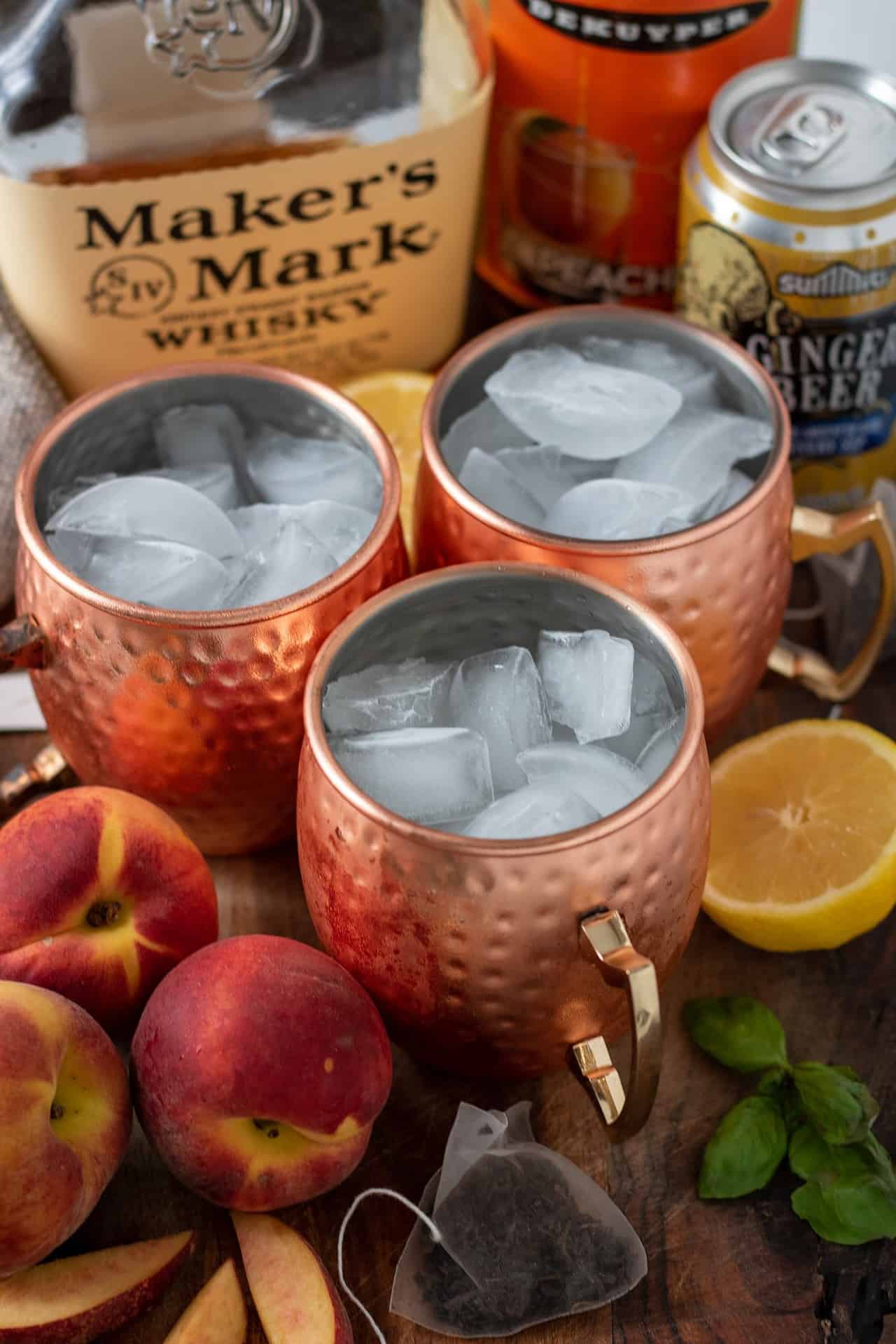 3 copper Moscow mule mugs filled with ice. There's peaches next to the mugs and a halved lemon. A bottle of bourbon is in the background. There's fresh mint sprinkled on the wooden surface too.