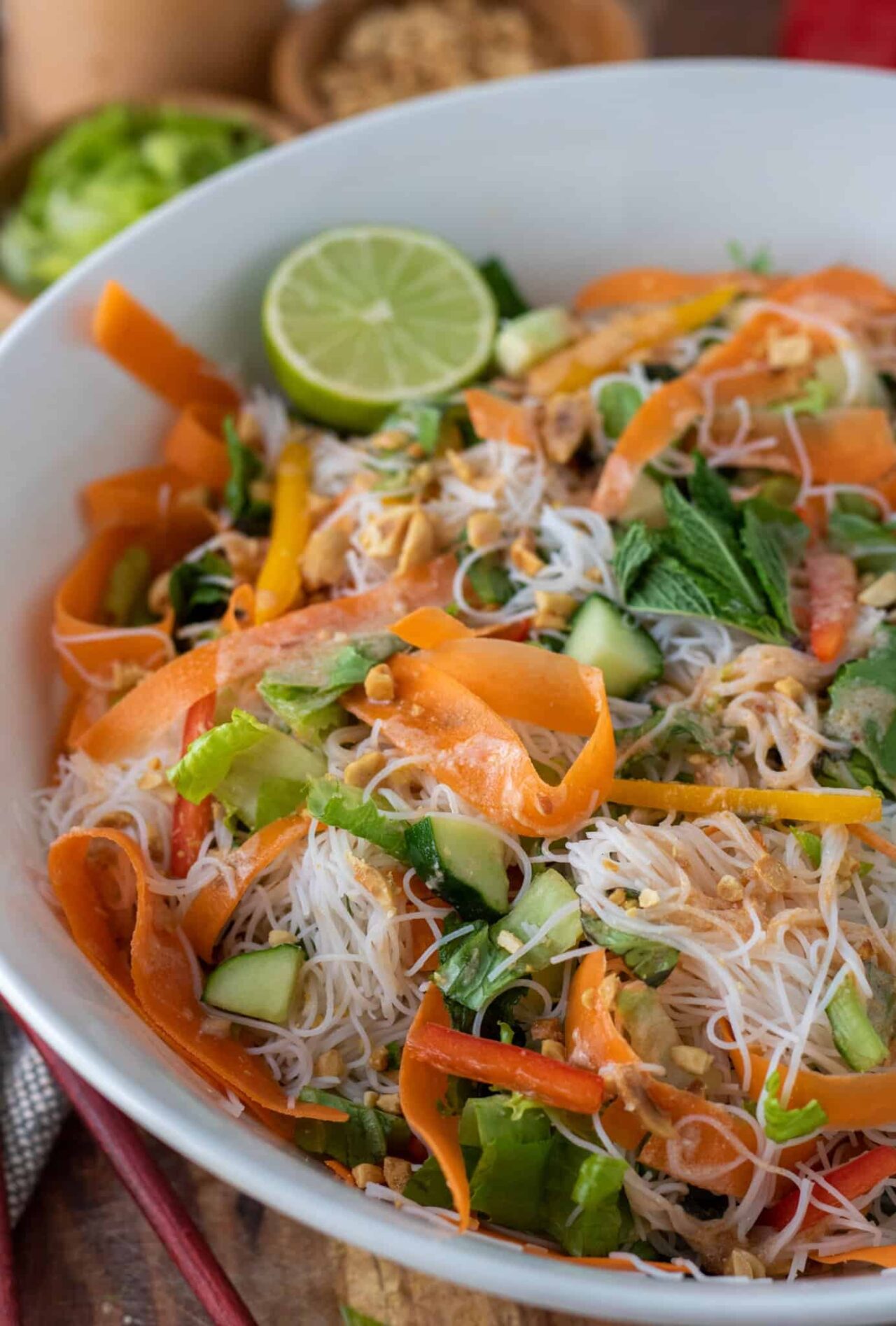 A large white bowl filled with cold noodle salad. There's half a lime on the edge of the bowl. It's garnished with crushed peanuts. The salad is bright and colorful.