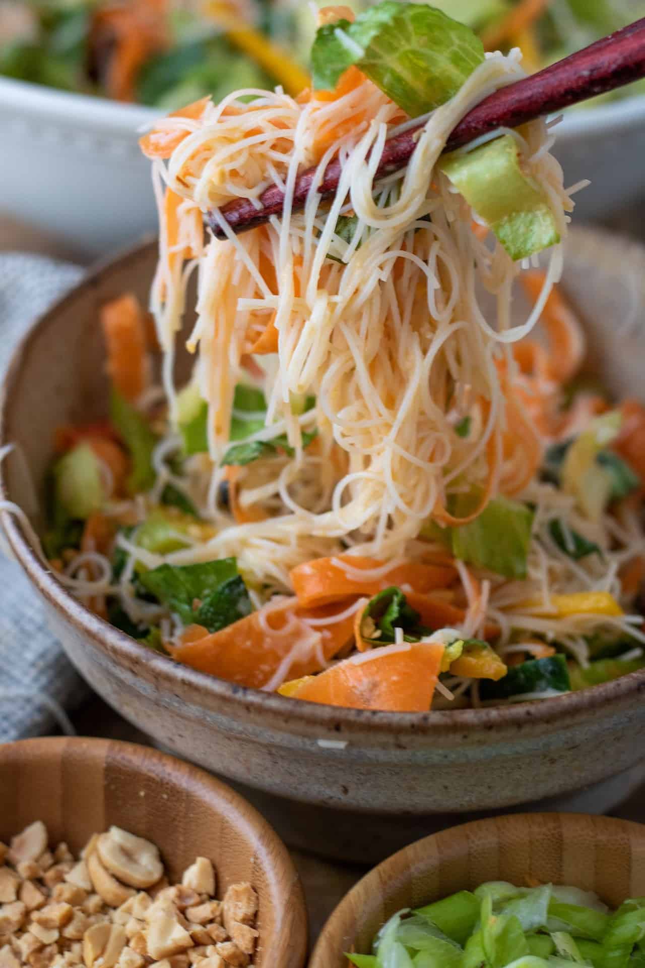 A brown speckled bowl filled with Asian noodle salad. Two chopsticks are holding up a mouthful of noodles. There's carrots, cucumbers and other veggies.