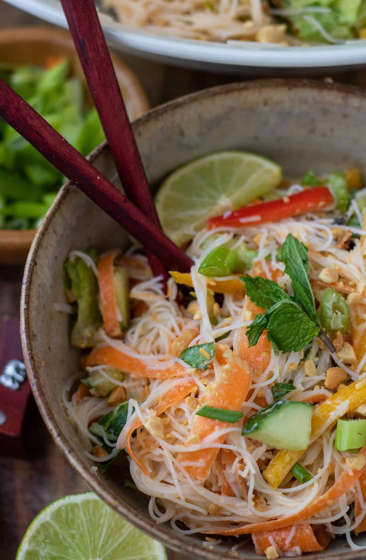A brown speckled bowl filled with Thai noodle salad. You can see the ribbons of carrots, diced cucumbers and sliced bell peppers. It's garnished with crushed peanuts and there's two chopsticks sticking out of the bowl