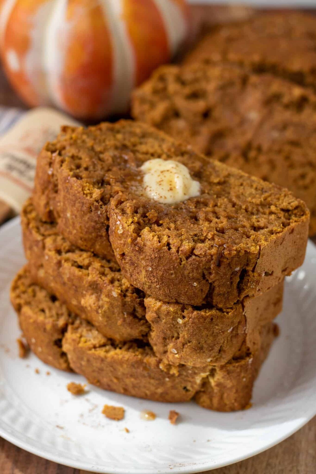 A small white plate topped with 3 thick slices of pumpkin banana bread. There's crumbs on the plate and the top piece of bread has a pad of melted butter on it. There's a small white and orange pumpkin in the background.