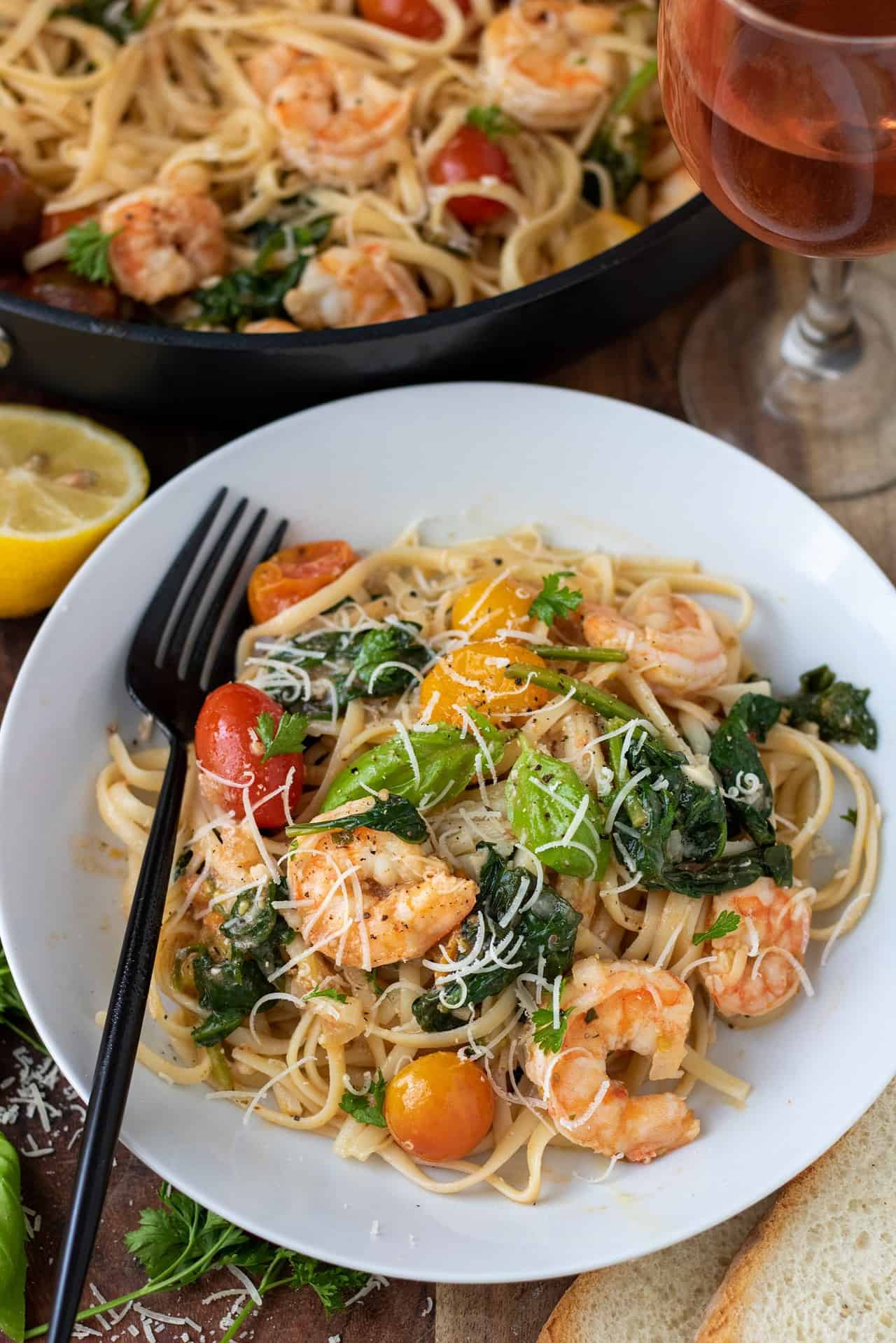 A white pasta bowl filled with linguine with shrimp, basil, tomatoes and spinach. There's a black fork resting on the dish. There's half a lemon, a glass of wine and crusty bread in the background.