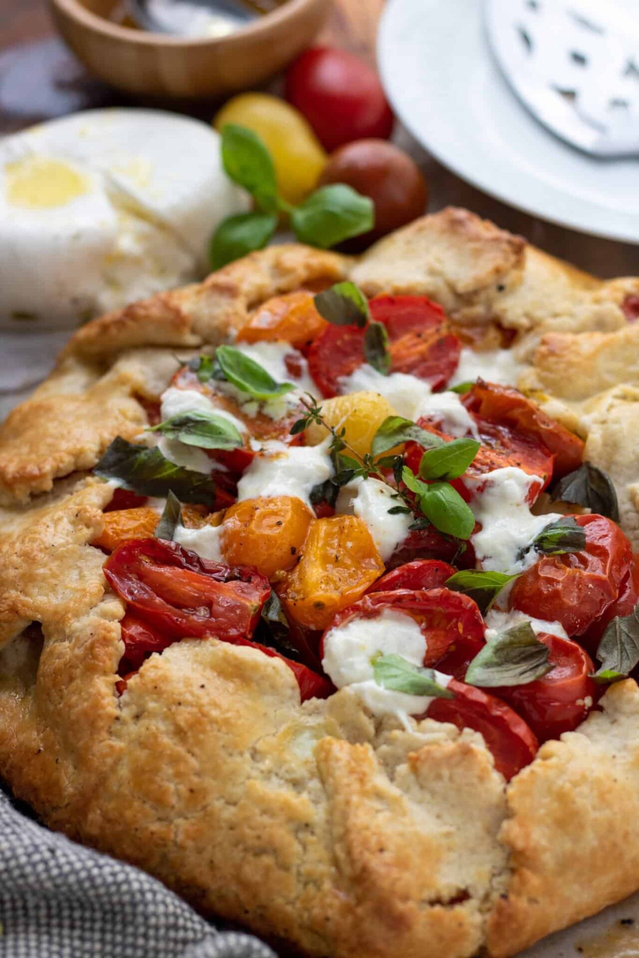 A savory tomato crostata topped with melted burrata cheese, fresh basil and a sprig of thyme. You can see a small white dish with a pie server in the background. You can also see a ball of fresh burrata cheese that's drizzled with olive oil