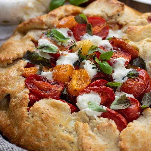 A savory tomato crostata topped with melted burrata cheese, fresh basil and a sprig of thyme. You can see a small white dish with a pie server in the background. You can also see a ball of fresh burrata cheese that's drizzled with olive oil