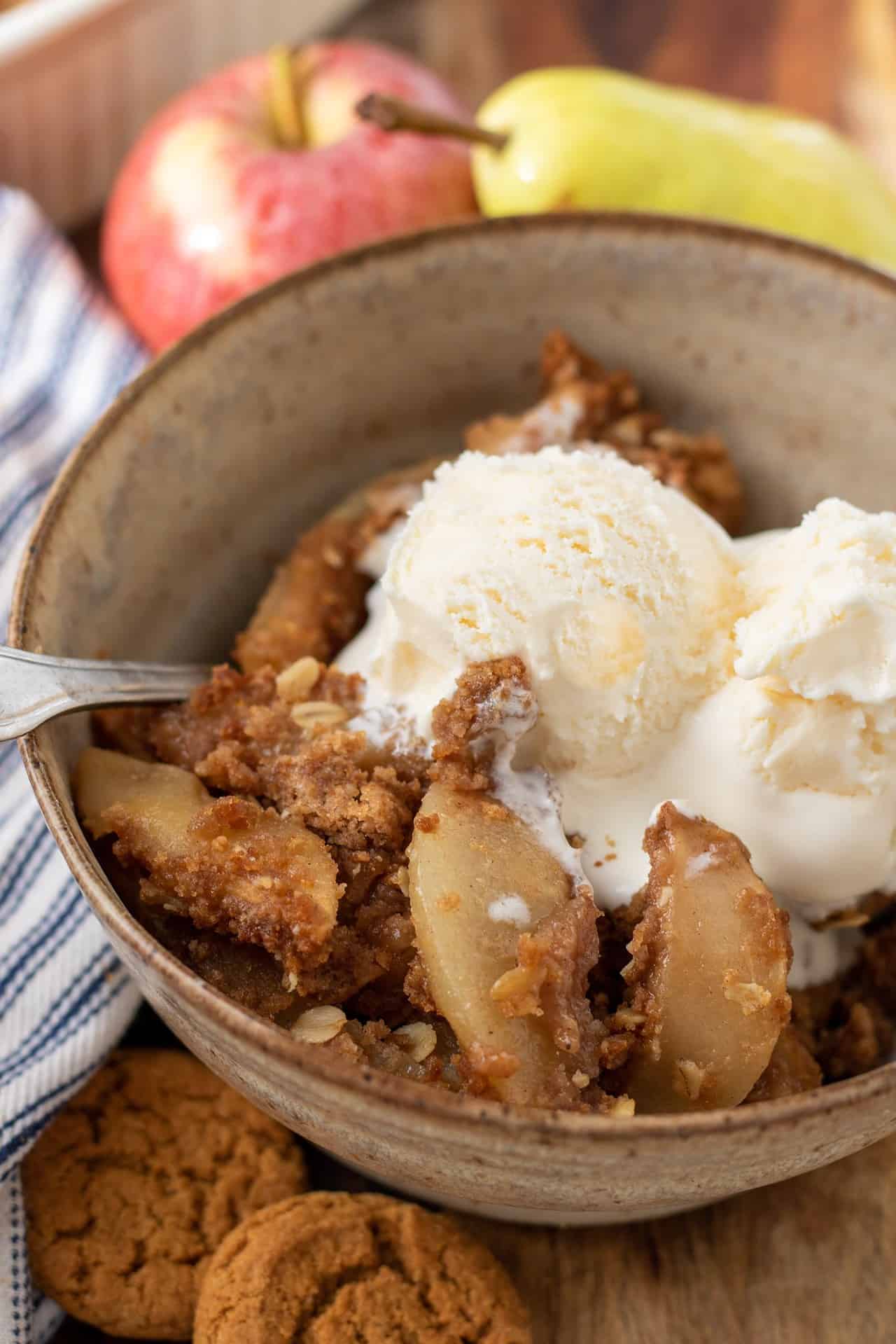 A brown speckled bowl with pear apple crisp that's topped with two scoops of vanilla ice cream. One of the scoops are slightly melted. There's a metal spoon sticking in the bowl. You can see two gingersnap cookies, a red apple and green pear in the background.
