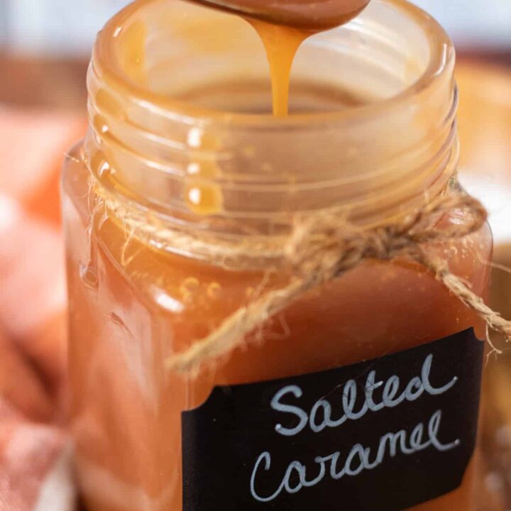 A small glass jar filled with salted caramel sauce. There's a spoon dipped into the sauce with the sauce dripping down. There's an orange and brown plaid dish towel next to the jar and sea salt sprinkled on the surface.