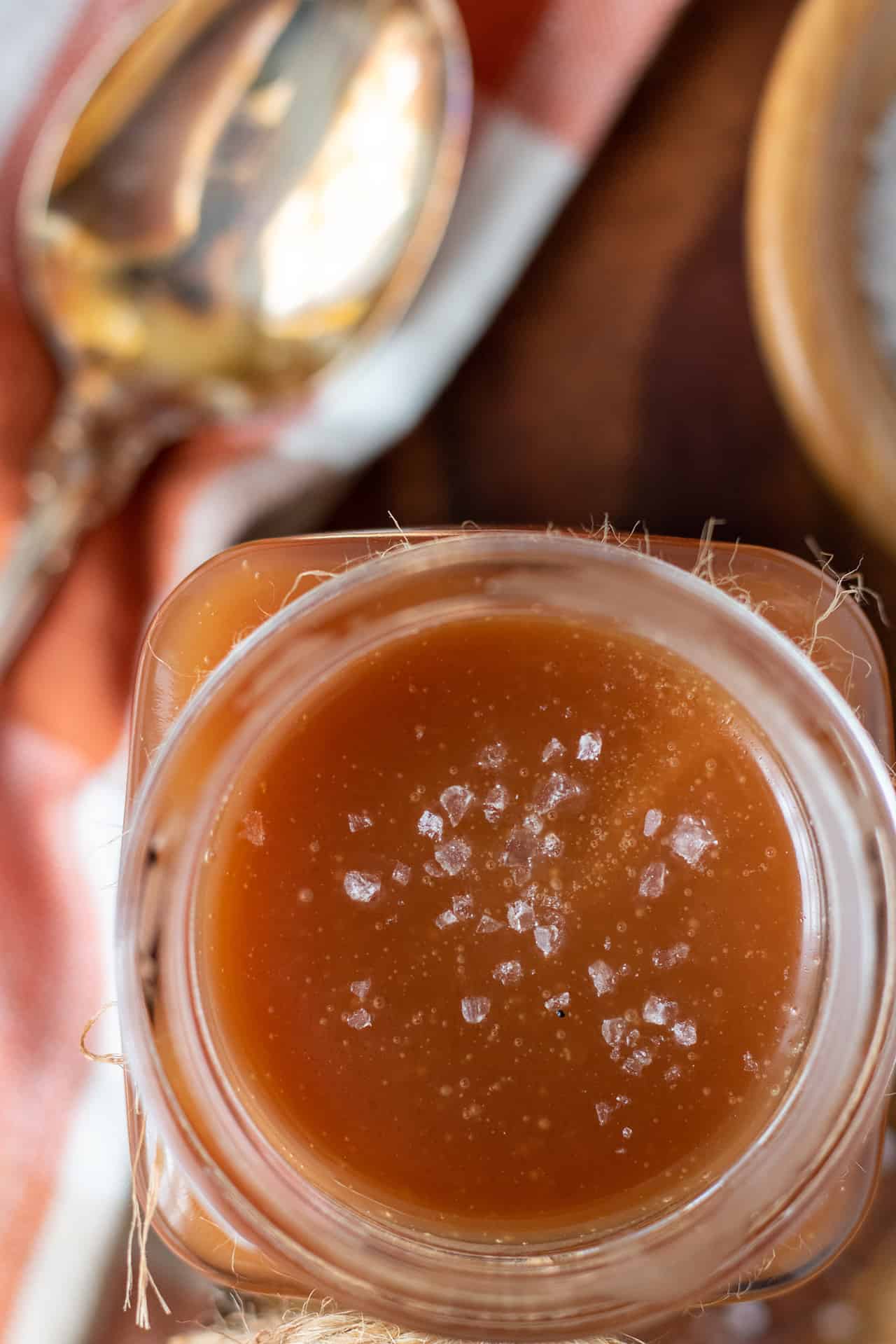 An overhead shot of a glass jar with caramel. There's sea salt chunks on top of it. There's a gold tarnished spoon in the background on a wooden surface.