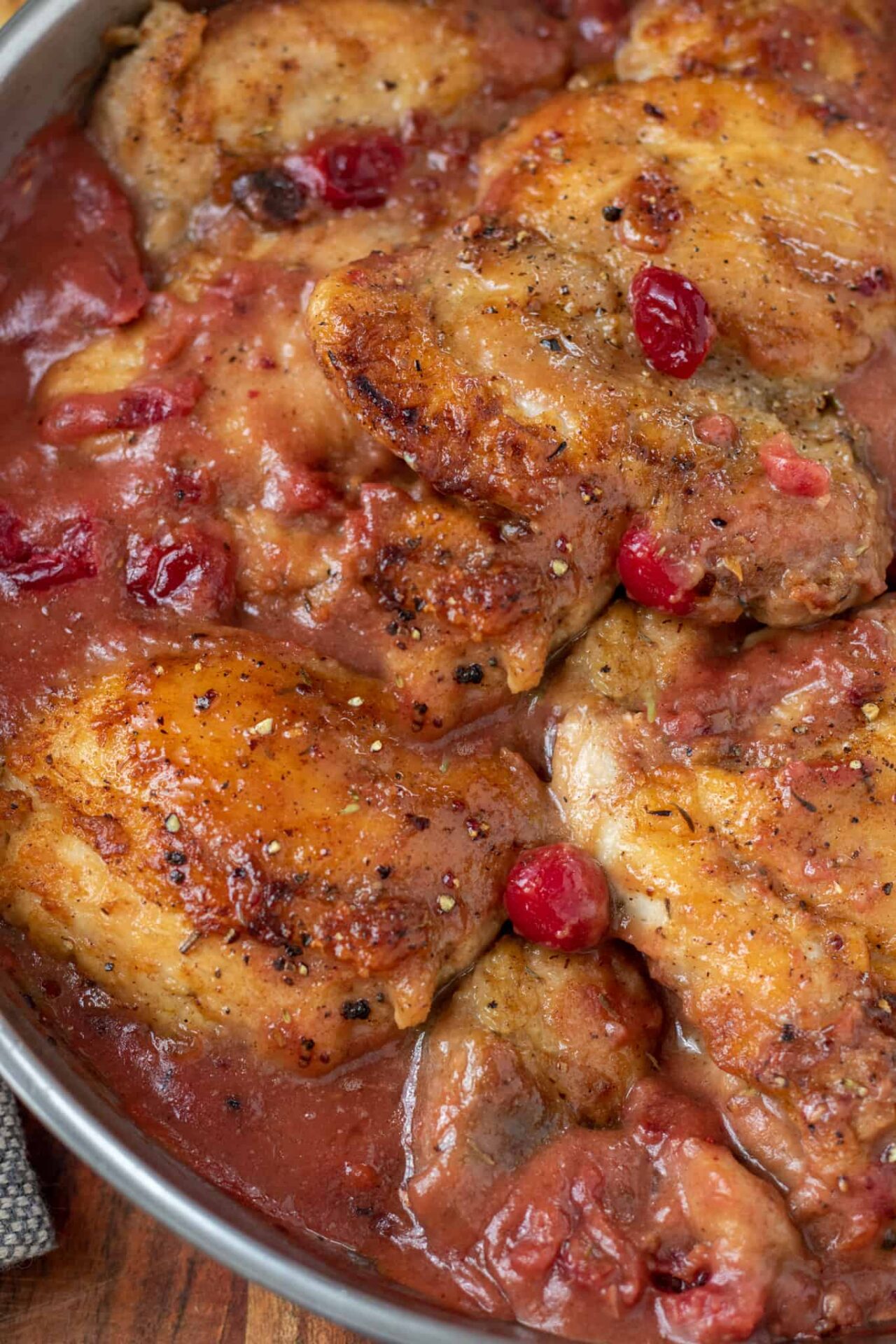 A skillet with golden brown pan seared chicken thighs in a cranberry gravy. There's whole cranberries in the sauce.